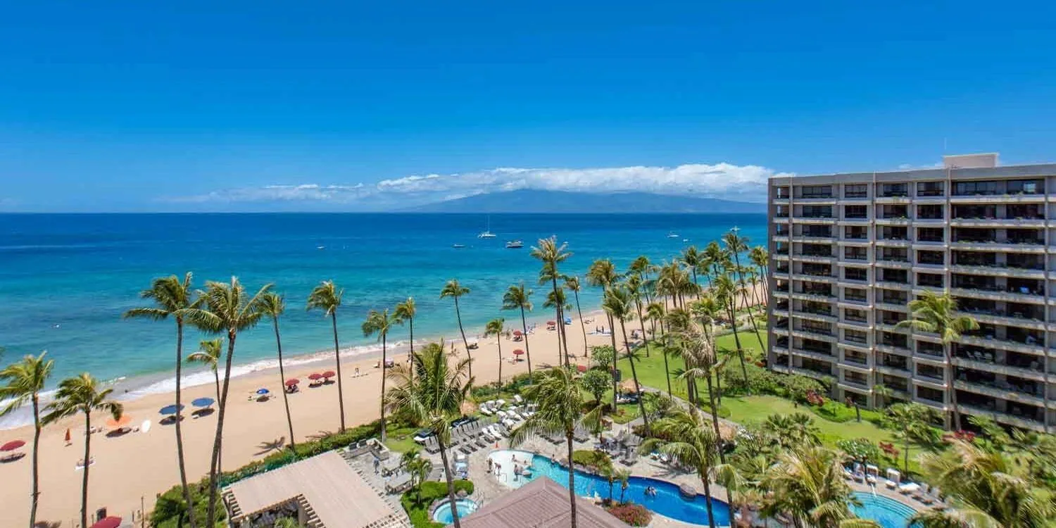 Kaʻanapali Beach from above: gold sand, calm turquoise water, a row of coconut palms, a neighbor island on the horizon, and an oceanfront resort condominium tower with lawns and pools.